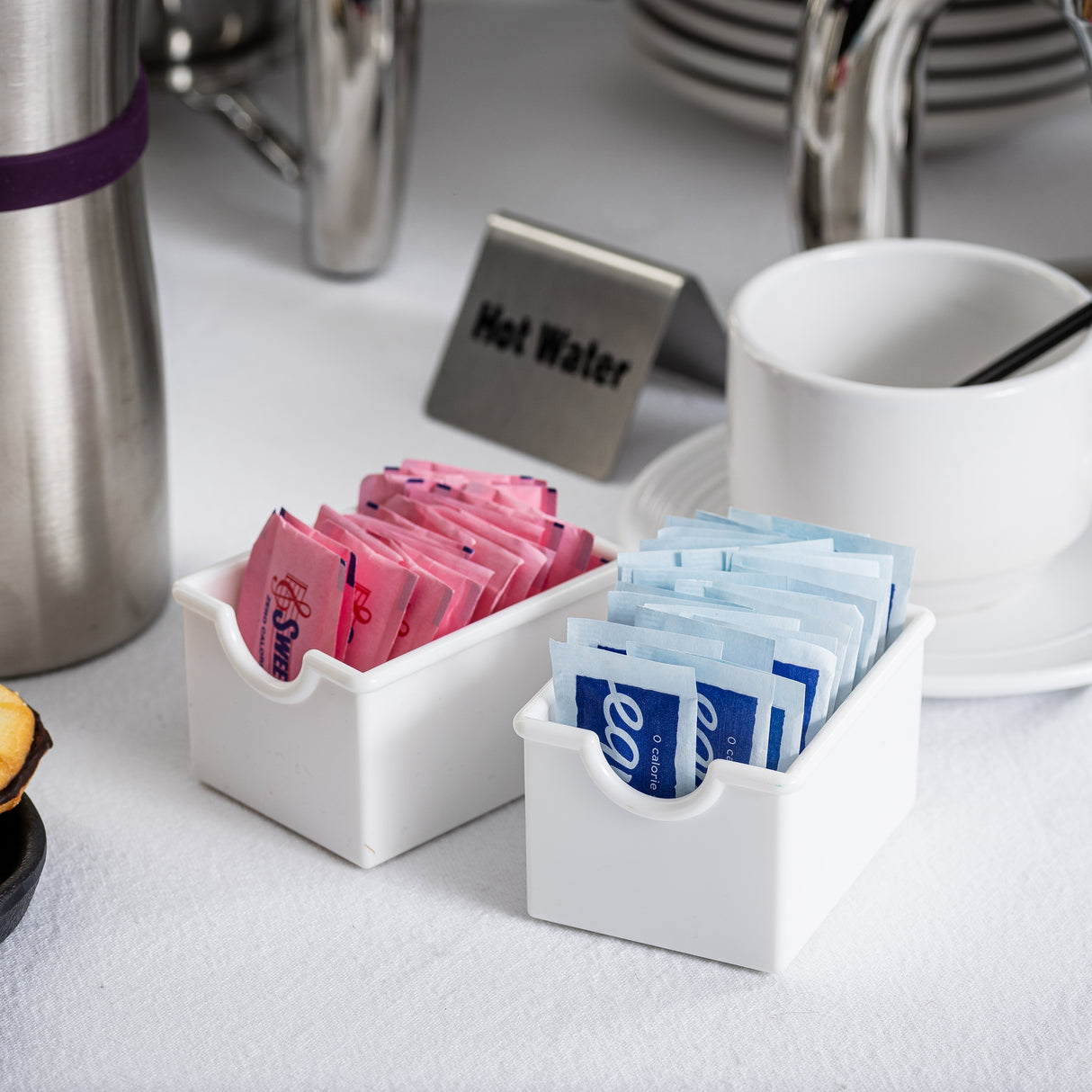 Two white, dishwasher safe containers serve as a stylish sugar packet rack for pink and blue sweetener packets. Nearby are stacked plates, a cup with a spoon, a metal thermos, and a “Hot Water” sign at this TableCraft beverage station.