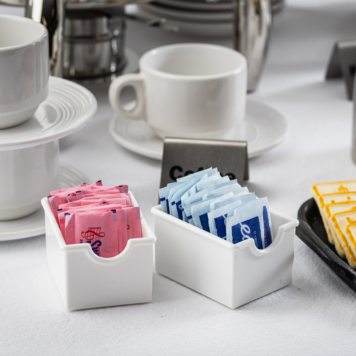 Two white containers act as a sugar packet rack, holding pink and blue sweetener packets on a white tablecloth. Stacked cups, a metal coffee pot, and a “Coffee” sign sit behind them. Slices of pound cake are arranged on a black plate to the right.