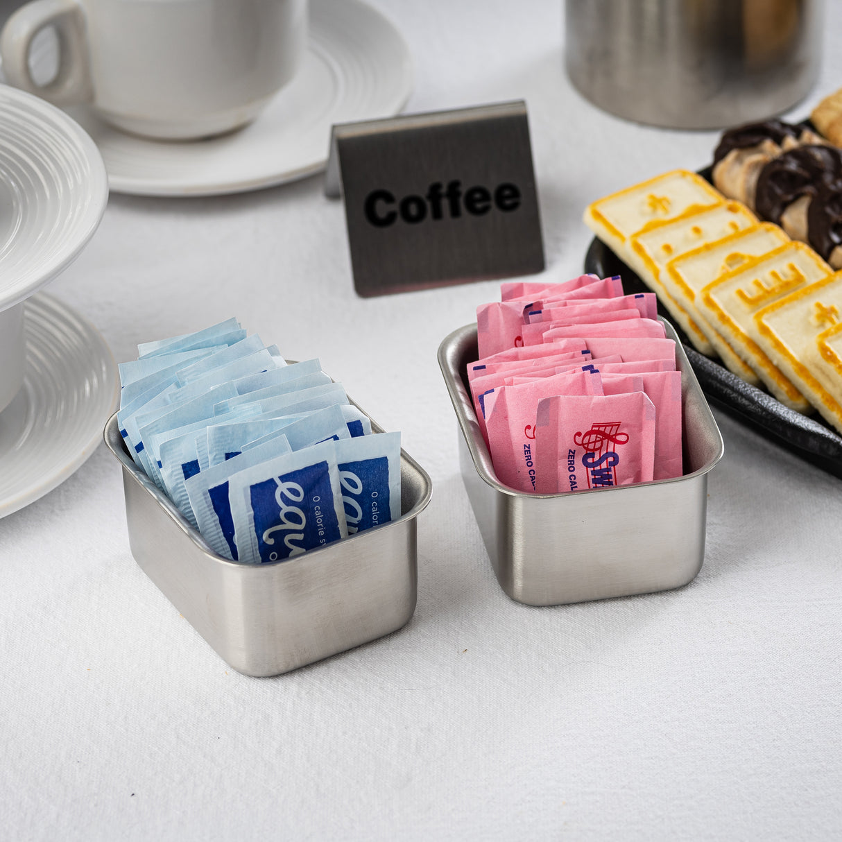 Two stainless steel containers hold blue and pink packets of artificial sweetener on a white tablecloth near plates, pastries, and a sign labeled “Coffee.” Part of a cup and saucer are also visible in the background.