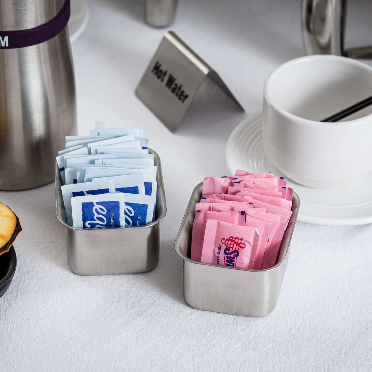Two stainless steel containers serve as a stylish sugar packet rack, holding blue Equal and pink Sweet’N Low packets. Nearby, a stacked white cup and saucer, small pastry, and “Hot Water” sign sit on a crisp white tablecloth.
