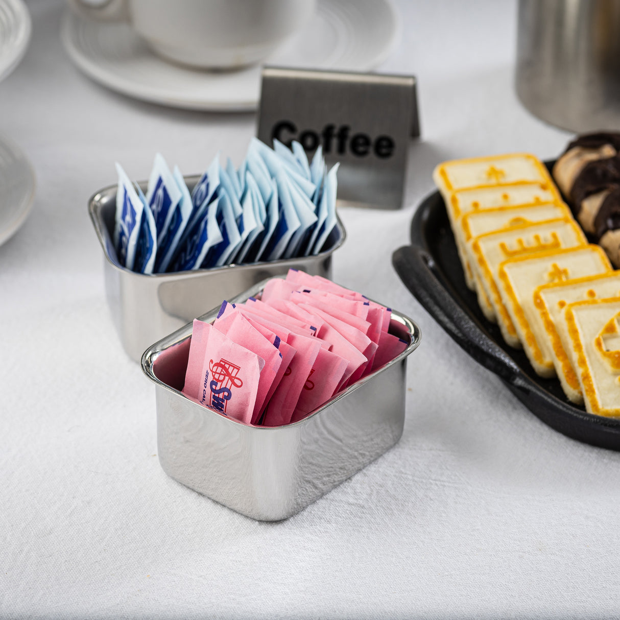 Two stainless steel containers hold pink and blue sugar or sweetener packets on a white tablecloth. Nearby are square cookies on a black tray and a small sign labeled Coffee. Porcelain cups and saucers are visible in the background.