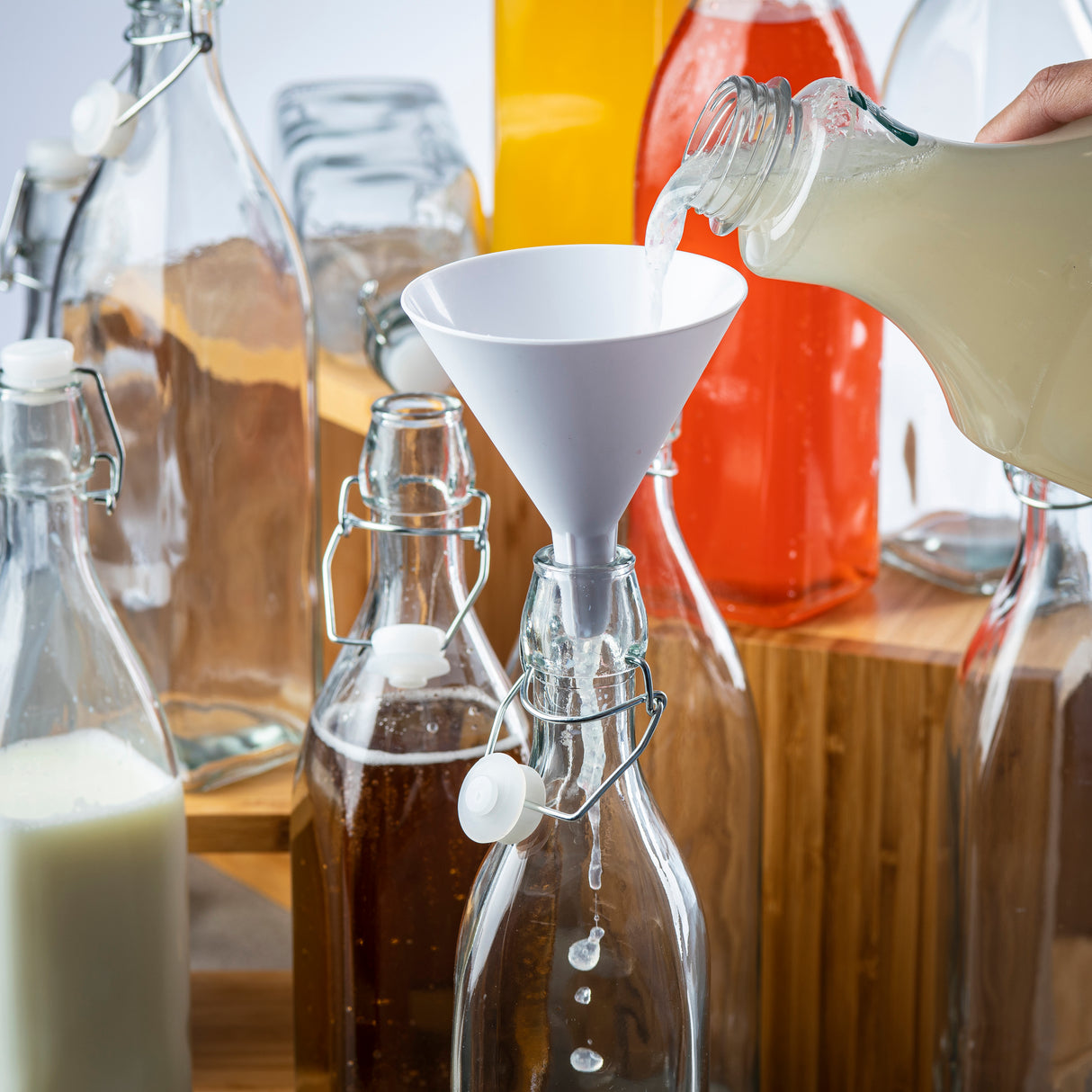 A hand pours a light-colored liquid from a jar into a glass bottle using a white plastic funnel. Surrounding the bottle are various dishwasher safe glass bottles filled with white, brown, and orange liquids, some with swing-top caps.