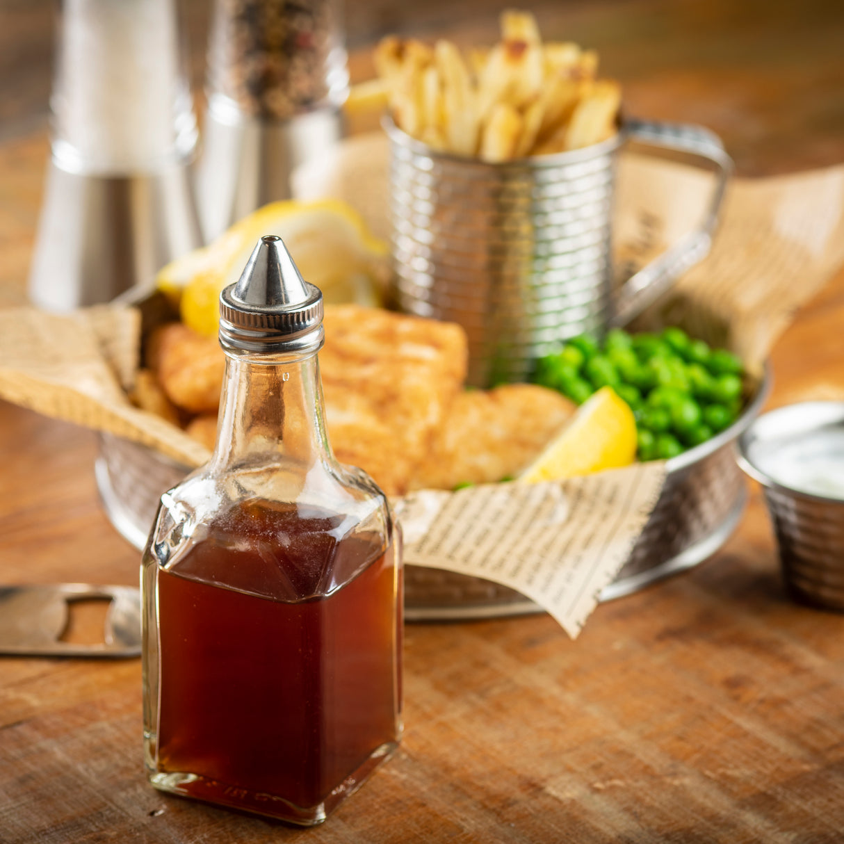 A glass vinegar dispenser with a stainless steel top sits in focus on a wooden table. In the background, fish and chips with mushy peas and tartar sauce are served on newspaper-style paper beside blurred salt and pepper shakers.