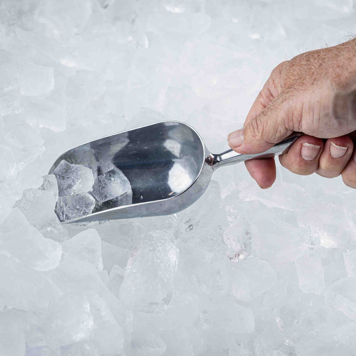A hand holds an aluminum Ice Scoop filled with ice cubes over a pile of loose ice. The background and surface are entirely covered with clear, irregularly shaped cubes for a cold, frosty appearance. The scoop has a 5 oz. capacity.