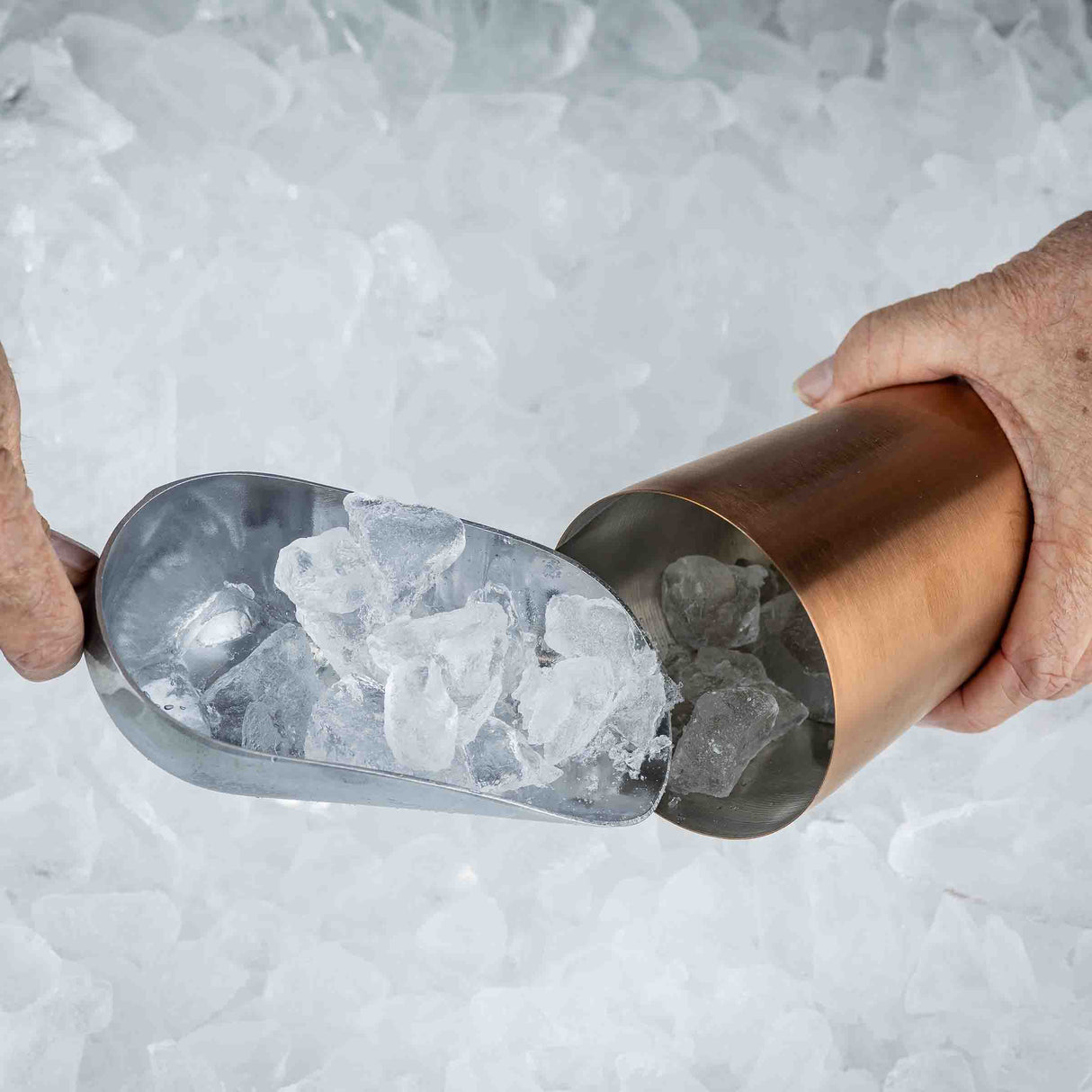 A close-up of hands using a 5 oz. aluminum Ice Scoop to pour ice cubes into a copper-colored cocktail shaker, with a background filled with crushed ice.