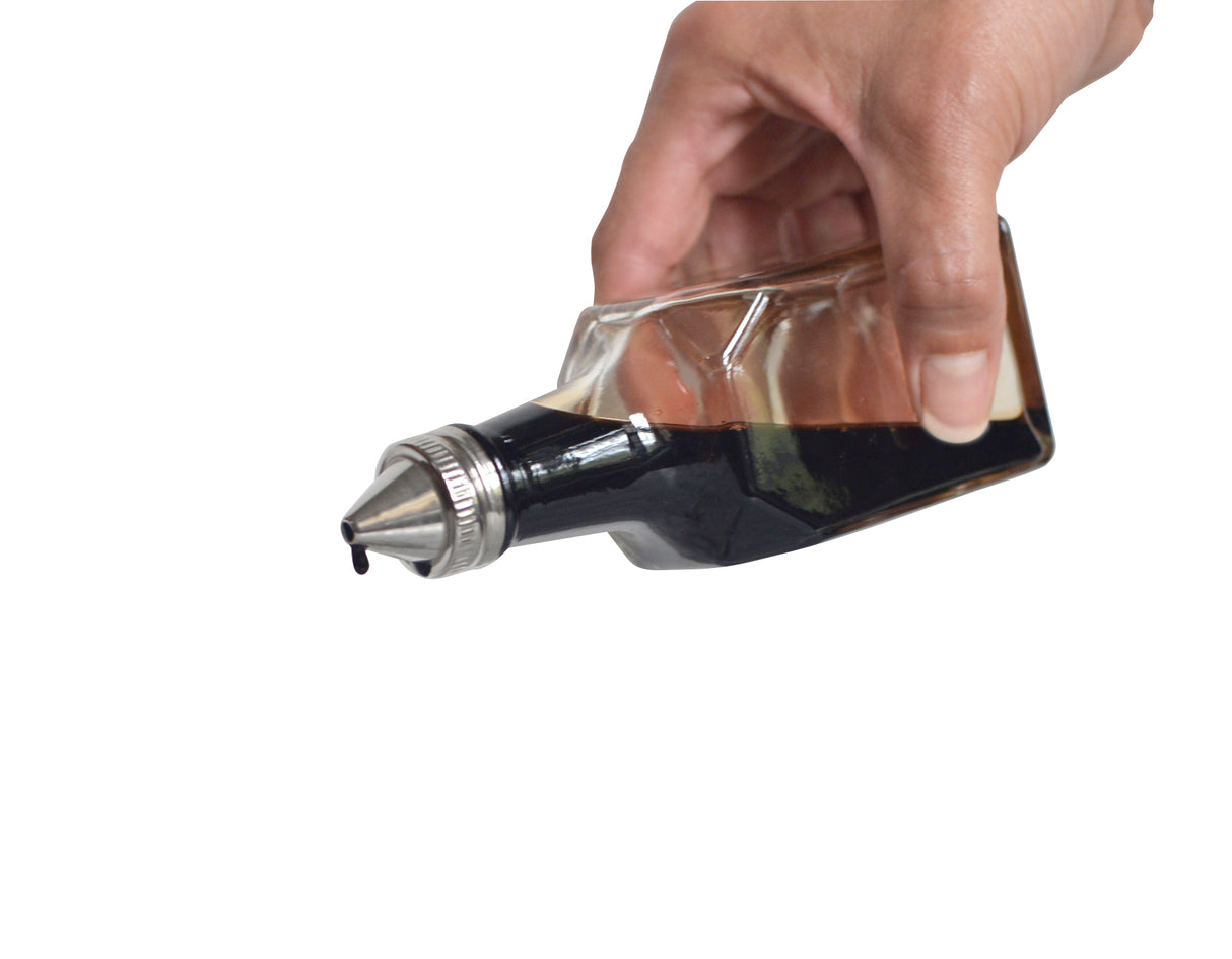 A hand tilts an 8 oz glass bottle Oil & Vinegar Dispenser with a metal spout, pouring a dark liquid, likely soy sauce, against a white background. A droplet hangs at the tip of the spout, about to fall.