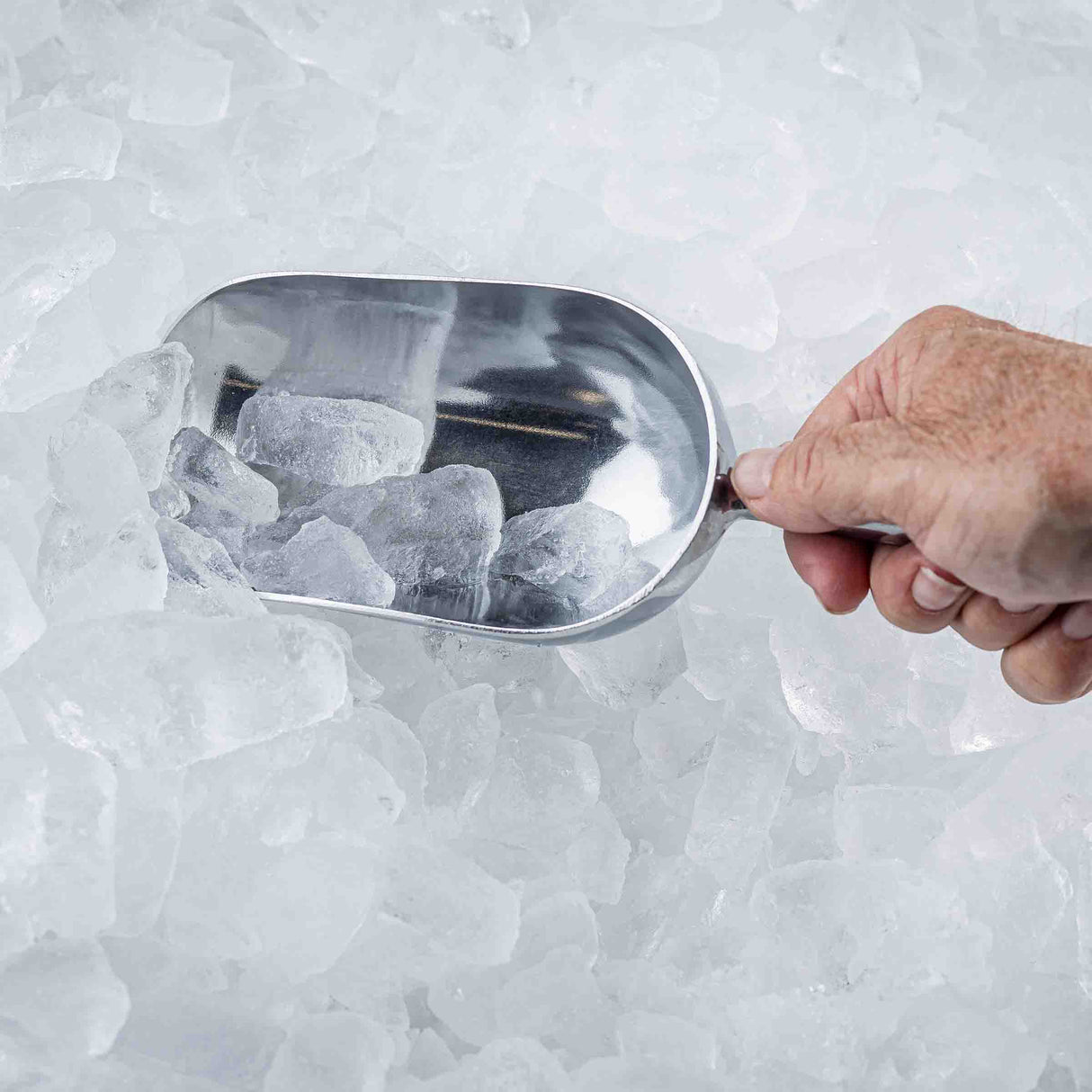 A hand holding a 12 oz. aluminum Ice Scoop picks up clear ice cubes from a pile. The background and surface are filled with irregular, white, translucent ice pieces, suggesting scooping ice from an ice bin or cooler.