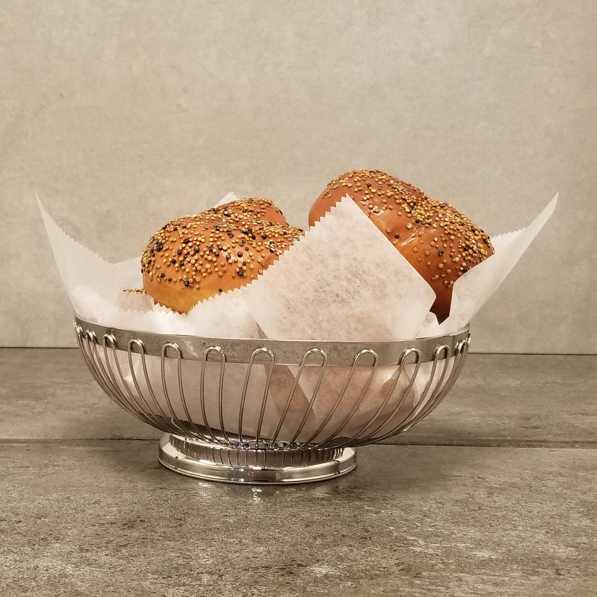 A stainless steel basket lined with white parchment paper holds two everything bagels topped with assorted seeds, sitting on a gray stone countertop against a plain gray wall.