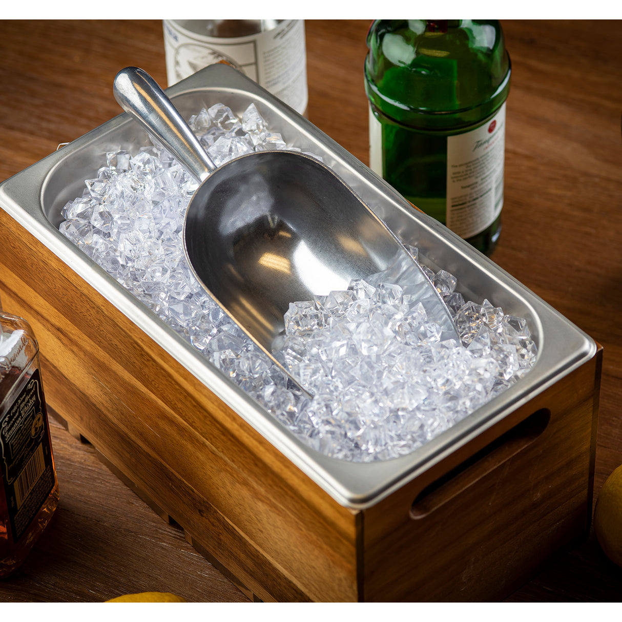 A 24 oz. aluminum Ice Scoop rests on clear ice cubes in a rectangular metal container set inside a wooden box. Bottles of alcohol and lemons sit on a wooden table in the background, creating a bar setup scene.
