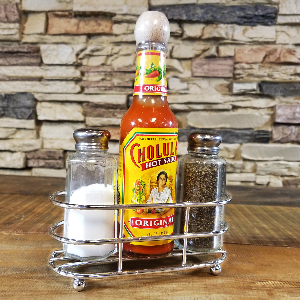 A chrome plated TableCraft condiment holder contains a bottle of Cholula hot sauce between clear glass shakers of salt and pepper. The background is a stone brick wall, and the set rests on a wooden surface.