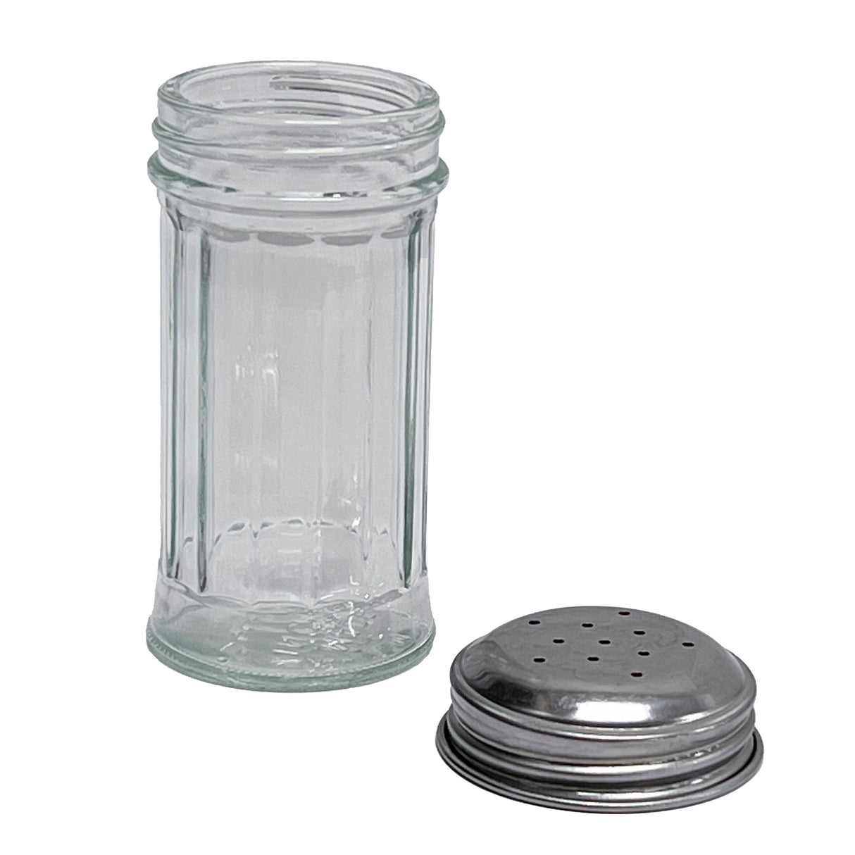 A clear, empty fluted glass salt/pepper shaker stands upright with vertical ridges along its sides. Its round, stainless steel top, featuring multiple small holes, is removed and placed on the white surface next to the shaker.