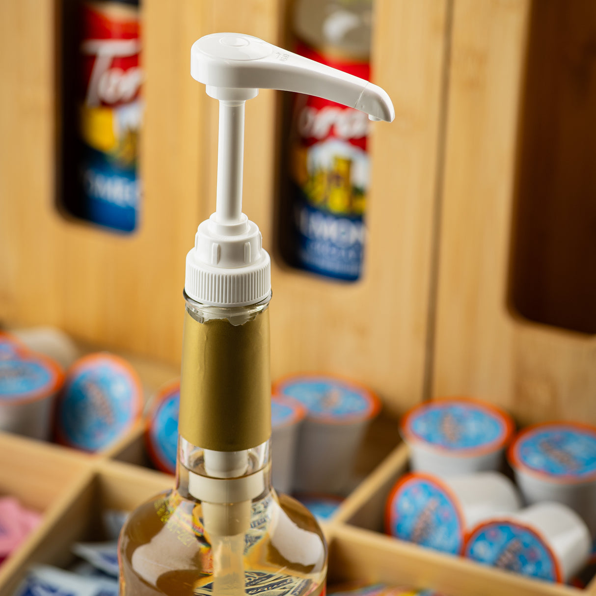 A close-up of a syrup bottle with a white, 1/4 oz syrup pump dispenser. In the background, wooden crates hold various syrup bottles and small coffee creamer cups, some with orange and blue lids—perfect for a busy coffee or beverage station.
