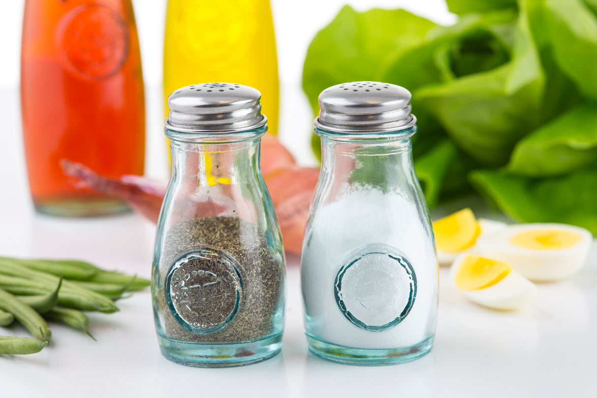Two glass shakers with stainless steel tops, one filled with ground black pepper and the other with salt, stand side by side. In the background are green vegetables, shallots, halved boiled eggs, and vibrant liquids on a white surface.