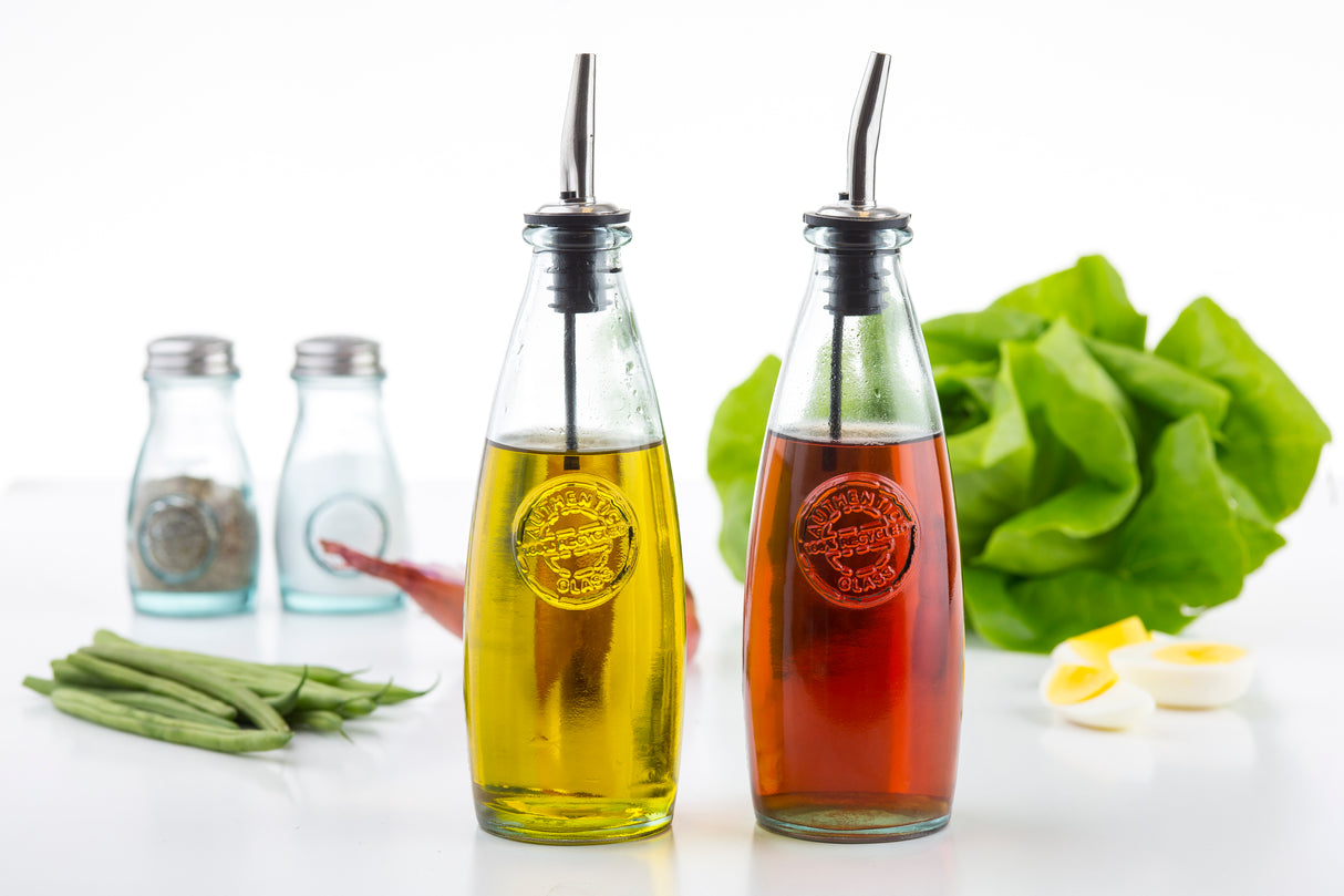 Two recycled glass Oil & Vinegar Dispensers with stainless steel tops hold yellow oil and red vinegar. Behind them sit blue salt and pepper shakers, leafy lettuce, green beans, a sliced boiled egg, and red onion on a white surface with a bright background.