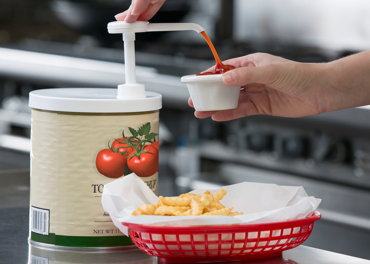 A hand uses the TableCraft 662K economy pump kit to dispense ketchup into a small cup. The large, tomato-labeled container sits behind a red basket of crinkle-cut French fries on a metal counter. The dispenser is dishwasher safe for easy cleaning.