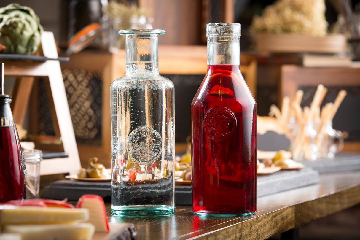 Two 41 oz glass carafes sit side by side on a table; one filled with sparkling clear water, the other with a deep red liquid. Both feature embossed emblems and are crafted from recycled glass, set against softly focused food and wooden décor.