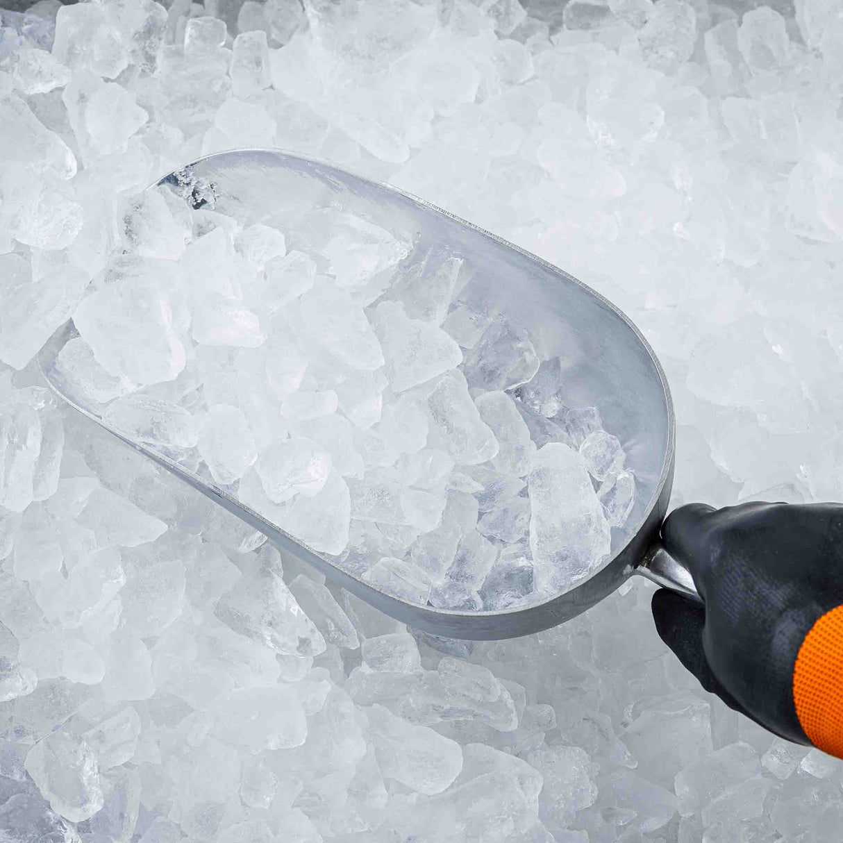 A gloved hand holds an 85 oz aluminum Ice Scoop filled with clear ice cubes, surrounded by more loose ice. The scoop is angled as if collecting the ice, and the background is entirely covered in irregularly shaped ice pieces.