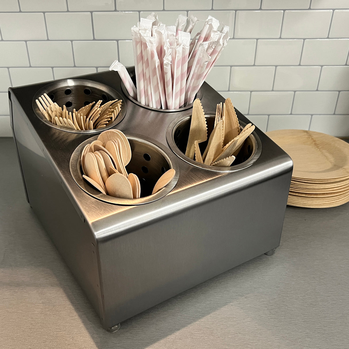 A stainless steel flatware cylinder holder contains wooden forks, spoons, knives, and striped paper straws. It sits on a gray countertop next to stacked round wooden plates, with a white subway tile backsplash in the background.
