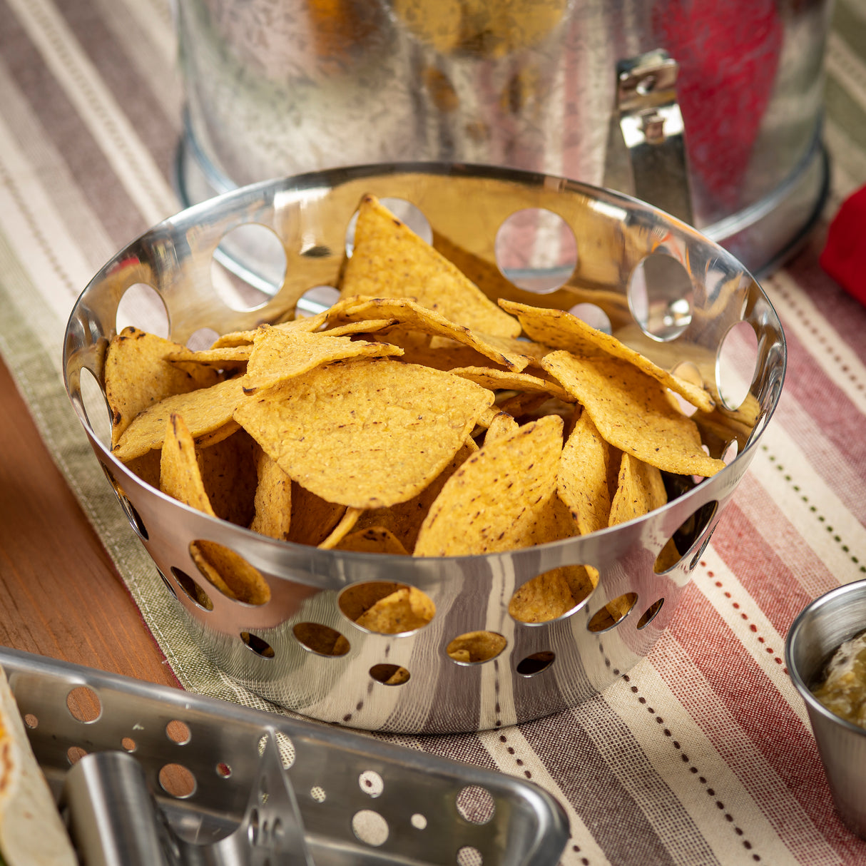 A stainless steel basket with round holes holds a pile of yellow corn tortilla chips. The bowl sits on a striped tablecloth in shades of beige, green, and red, with metallic dishes and utensils partially visible nearby.