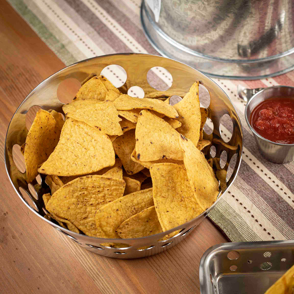 A stainless steel bowl filled with yellow tortilla chips sits on a wooden table next to a small cup of red salsa. The table is covered with a striped, earth-toned runner, and part of a metal tray and covered dish are also visible nearby.