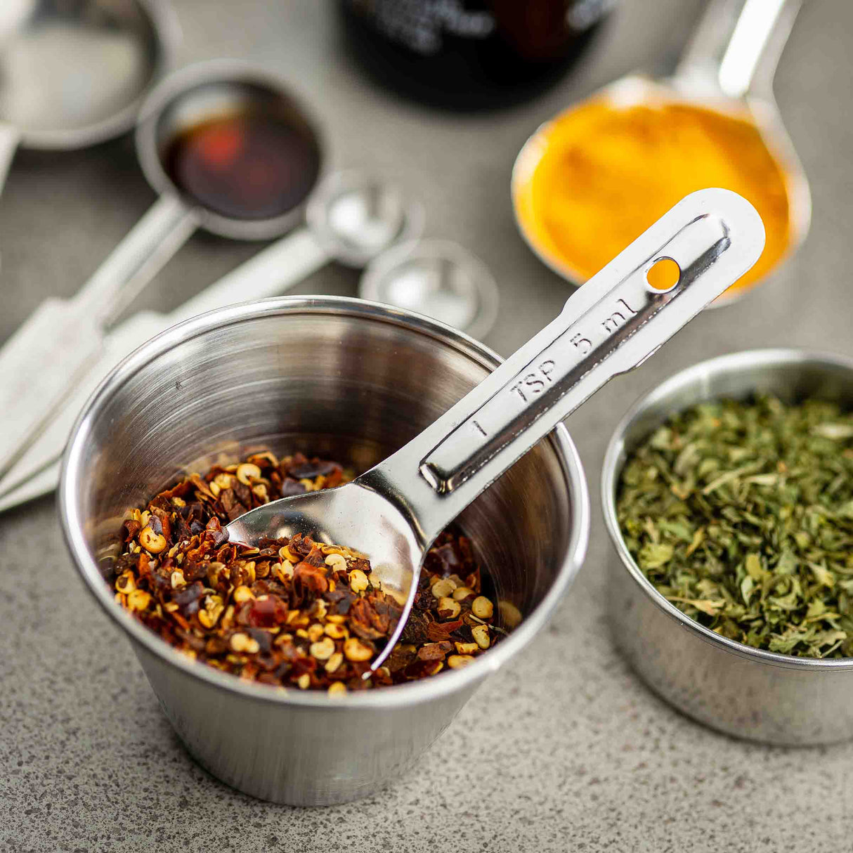 A stainless steel measuring spoon labeled 1 tsp rests in a cup filled with crushed red pepper flakes. Nearby, metal cups hold dried herbs and bright yellow turmeric powder, plus assorted measuring spoons—all on a gray countertop.