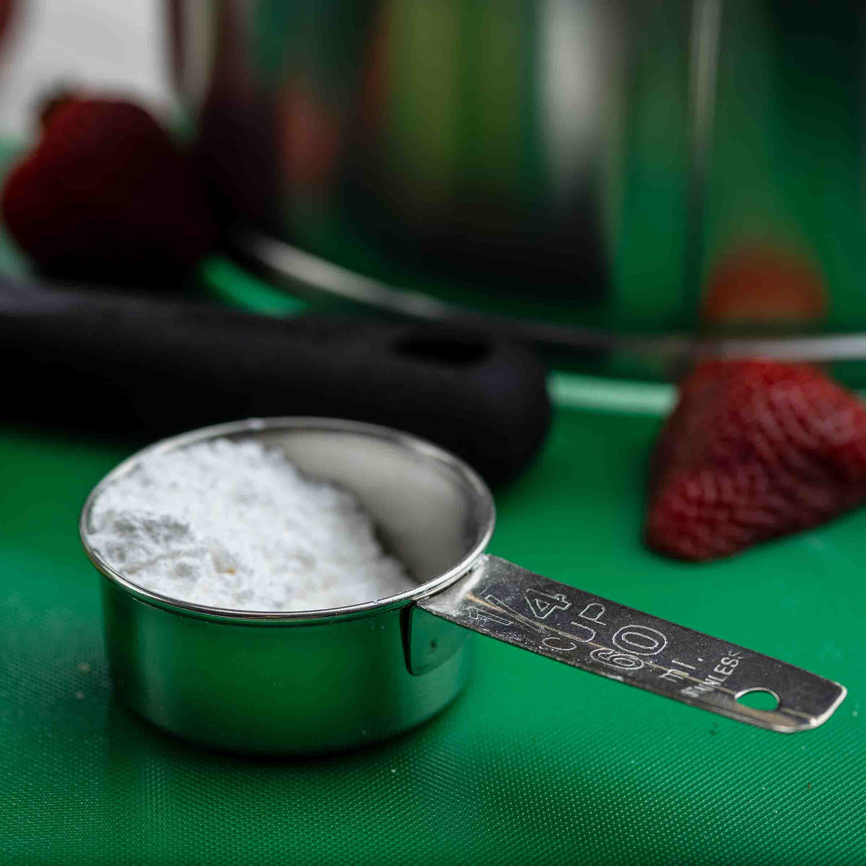 A stainless steel Measuring Cup filled with powdered sugar sits on a green surface. Blurry strawberries, a black utensil, and a stainless steel bowl appear in the background. The 1/4 cup measurement is marked on the cup’s handle.