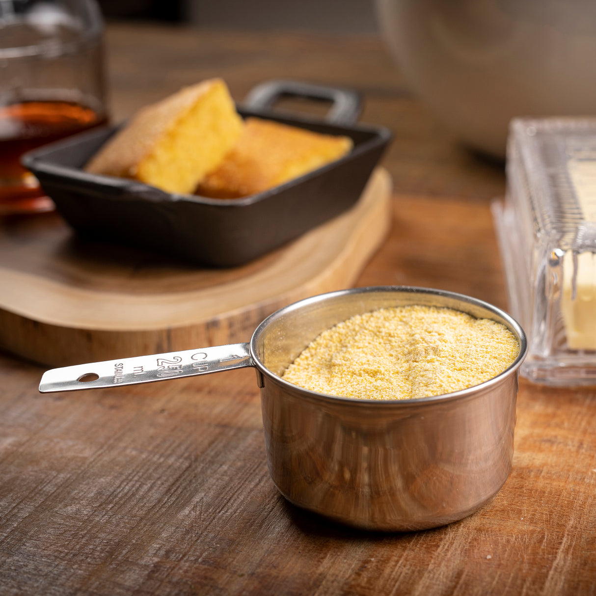 A stainless steel measuring cup filled with cornmeal sits on a wooden table. In the background, a small cast iron dish with cornbread, a stick of butter in a butter dish, and a glass with brown liquid are all slightly out of focus.