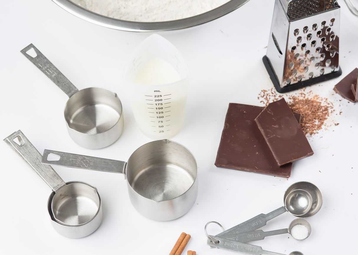 A flat lay of baking tools and ingredients: three stainless steel measuring cups, nested metal measuring spoons, a clear dishwasher safe measuring cup with liquid, dark chocolate bars, a box grater with chocolate shavings, cinnamon sticks, and flour—all on a white surface.