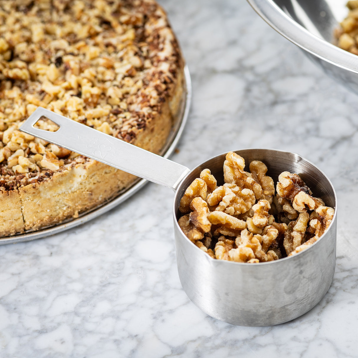 A stainless steel measuring cup filled with shelled walnut halves sits on a marble surface. In the background, a round cake topped with chopped walnuts and a partially visible metal bowl complete the scene.