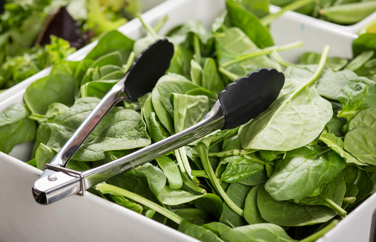A pair of stainless steel tongs rests on a pile of fresh spinach leaves in a white container, surrounded by other leafy greens. The crisp, vibrant spinach is ready for serving with these handy, dishwasher safe tongs.