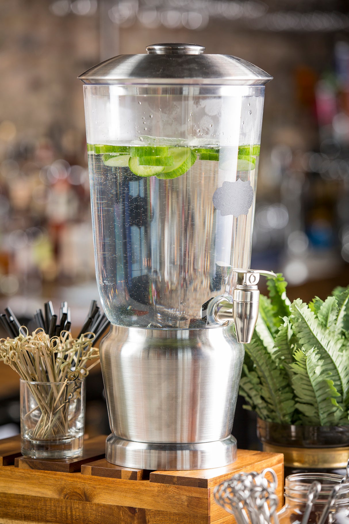 A large BPA free stainless steel beverage dispenser filled with water and floating lime slices sits on a wooden stand. Nearby are drink stirrers, metal tongs, and a green fern, with a blurred bar or café setting in the background.