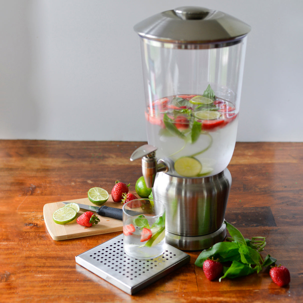 A BPA free glass beverage dispenser filled with water, sliced limes, strawberries, and mint sits on a wooden table. In front, a glass with the same ingredients, a metal drip tray, and a cutting board with limes and strawberries are visible.