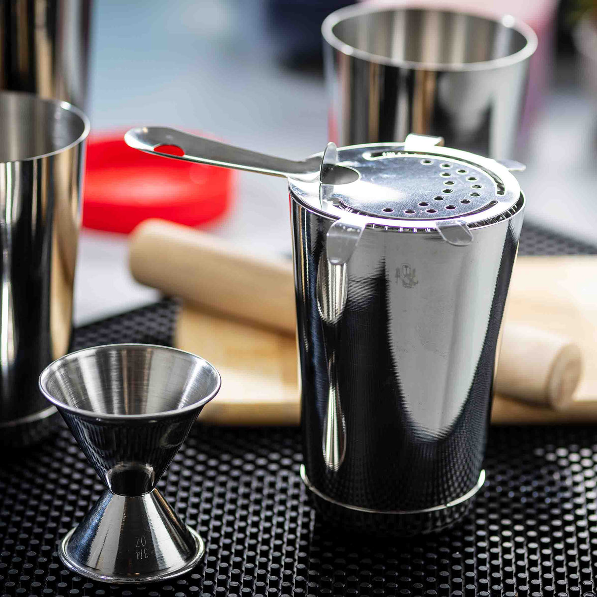 A close-up of shiny stainless steel bar tools on a black mat: a dishwasher safe Bar Shaker with strainer, double-sided jigger, muddler, and another metal cup in the background, all with blurred colorful objects behind them.