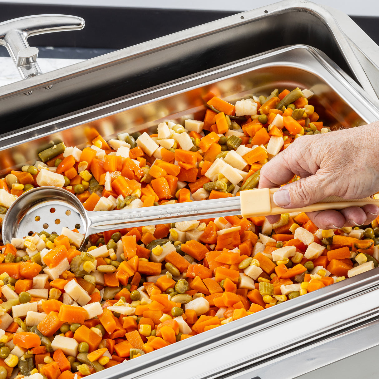 A hand uses a perforated serving spoonout to scoop up a mix of diced cooked vegetables, including carrots, green beans, and parsnips, from a stainless steel buffet tray.