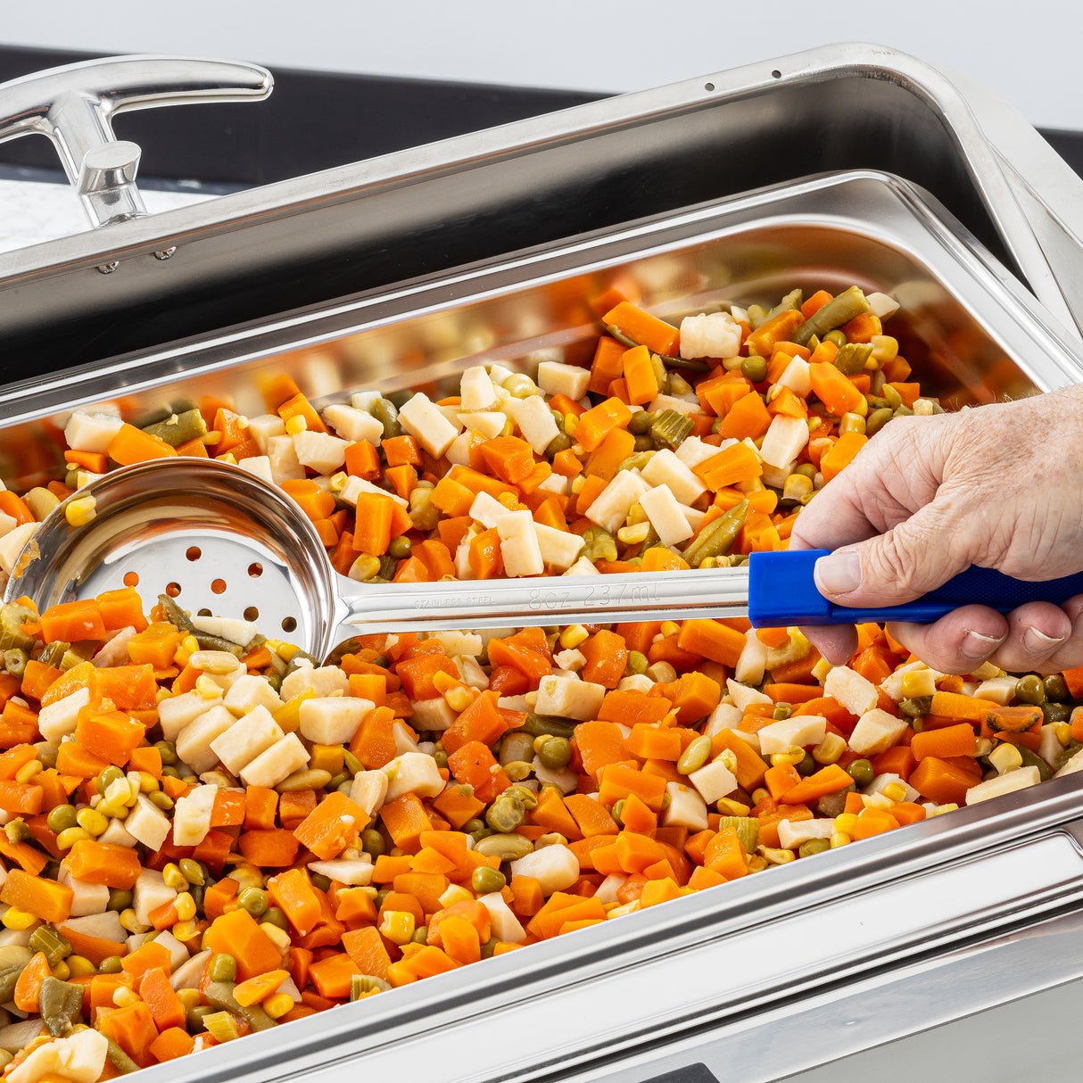 A close-up of a hand holding the Spoonout slotted serving spoon with a blue handle, scooping a colorful mix of diced veggies from a stainless steel food warmer tray; the Spoonout is durable and dishwasher safe.
