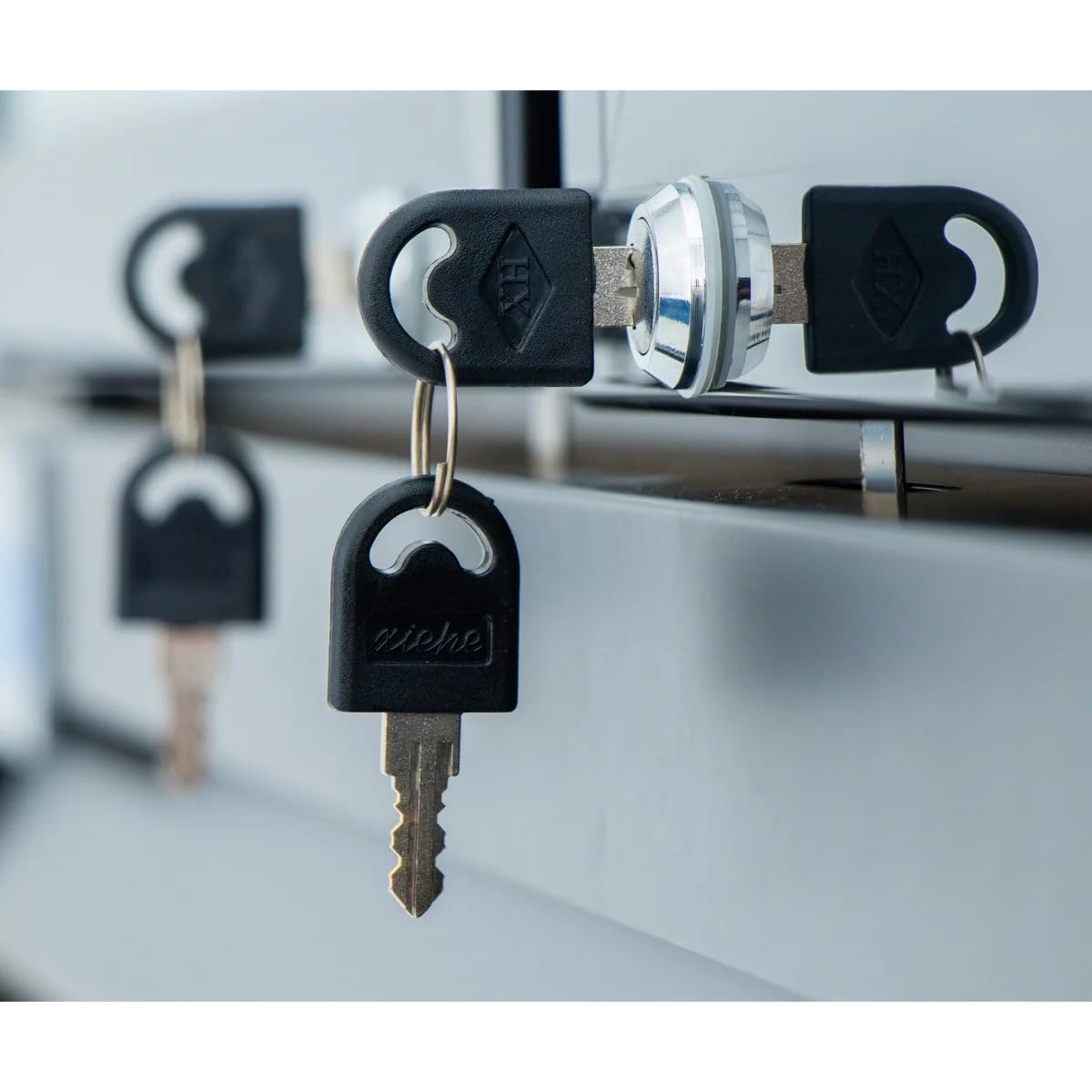 Close-up of three black keys inserted into silver locks on a row of metallic commercial refrigeration drawers. The keys have circular holes in the handles and one features a keychain labeled eische. Focus is on the middle key; background softly blurred.