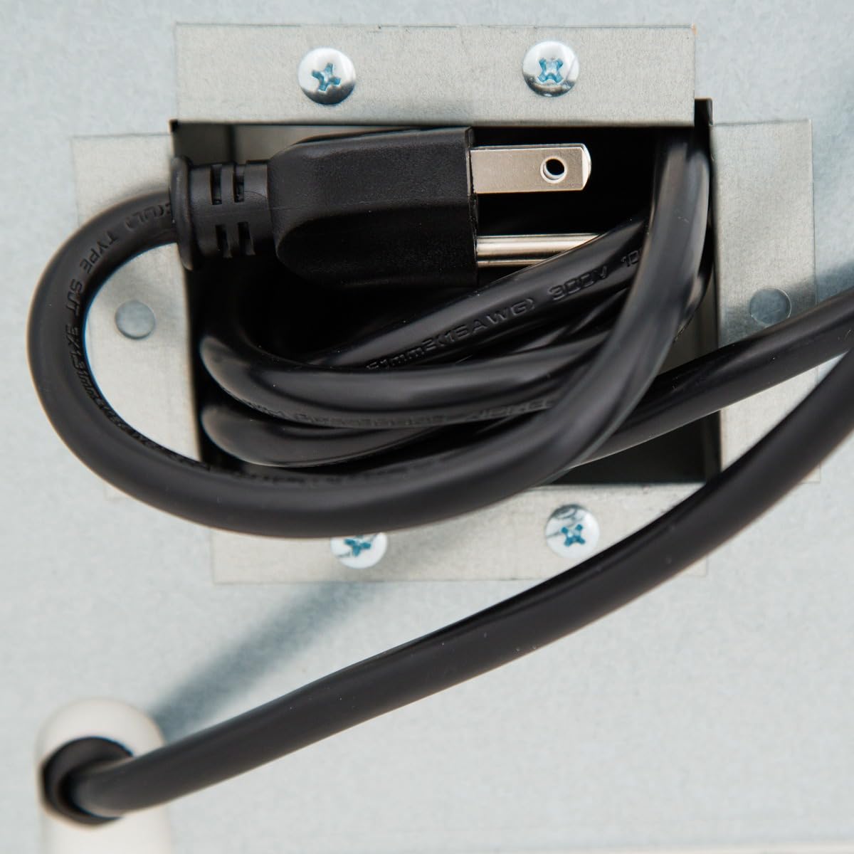 A black electrical plug and its coiled cord are neatly stored inside a metal box, secured with four screws at the corners—typical of a commercial refrigerator. Another black cord enters the box from the bottom left, both against a metallic surface.