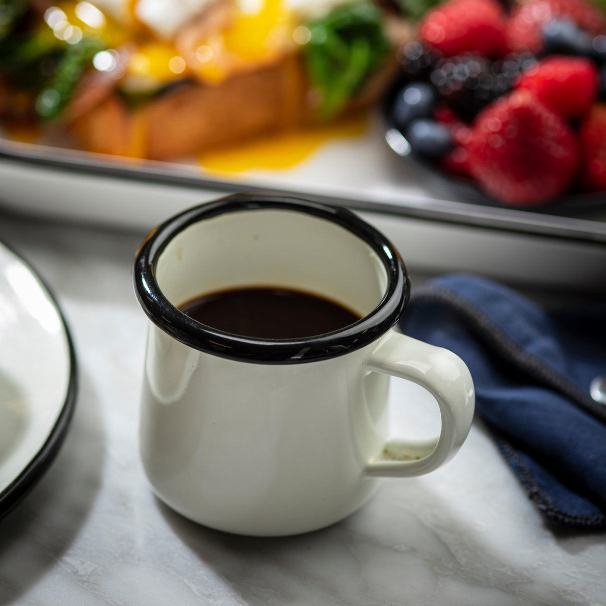 A white porcelain enamel mug with a black rim filled with black coffee sits on a marble surface. In the blurred background, there's a plate with berries and another with toast topped with greens and a runny egg, plus a blue napkin and spoon nearby.