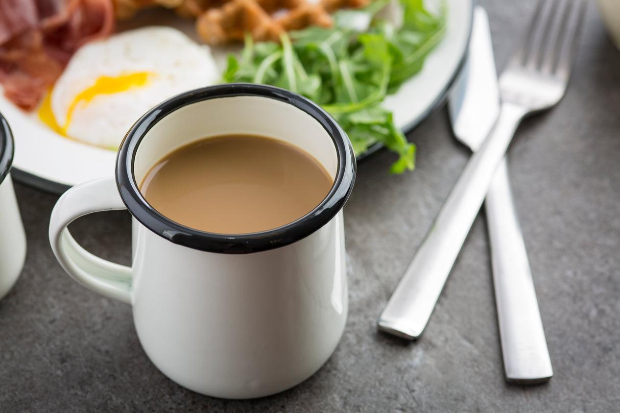 A white enamelware mug with a black rim filled with coffee sits on a gray surface. Behind it is a plate holding a sunny-side-up egg, leafy greens, and other breakfast items. A knife and fork rest beside the dishwasher safe mug.