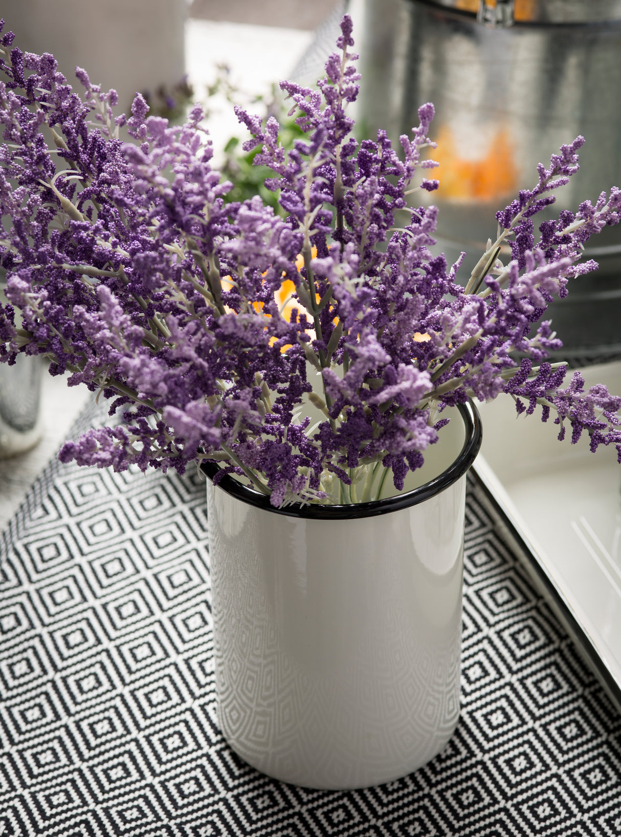 A stackable white ceramic cup with a black rim holds a bouquet of artificial lavender flowers. The cup sits on a black-and-white geometric-patterned placemat, with a softly blurred background hinting at other objects.