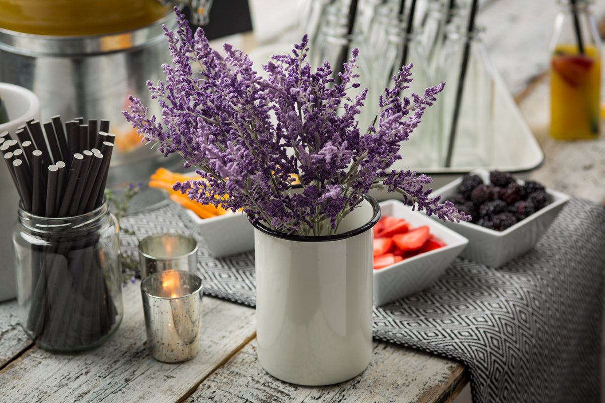 A white enamelware side cup holds a bouquet of purple lavender on a rustic wooden table. Nearby are black straws in a jar, lit candle holders, and white bowls with cut fruit and berries. A gray patterned cloth and glassware are in the background.