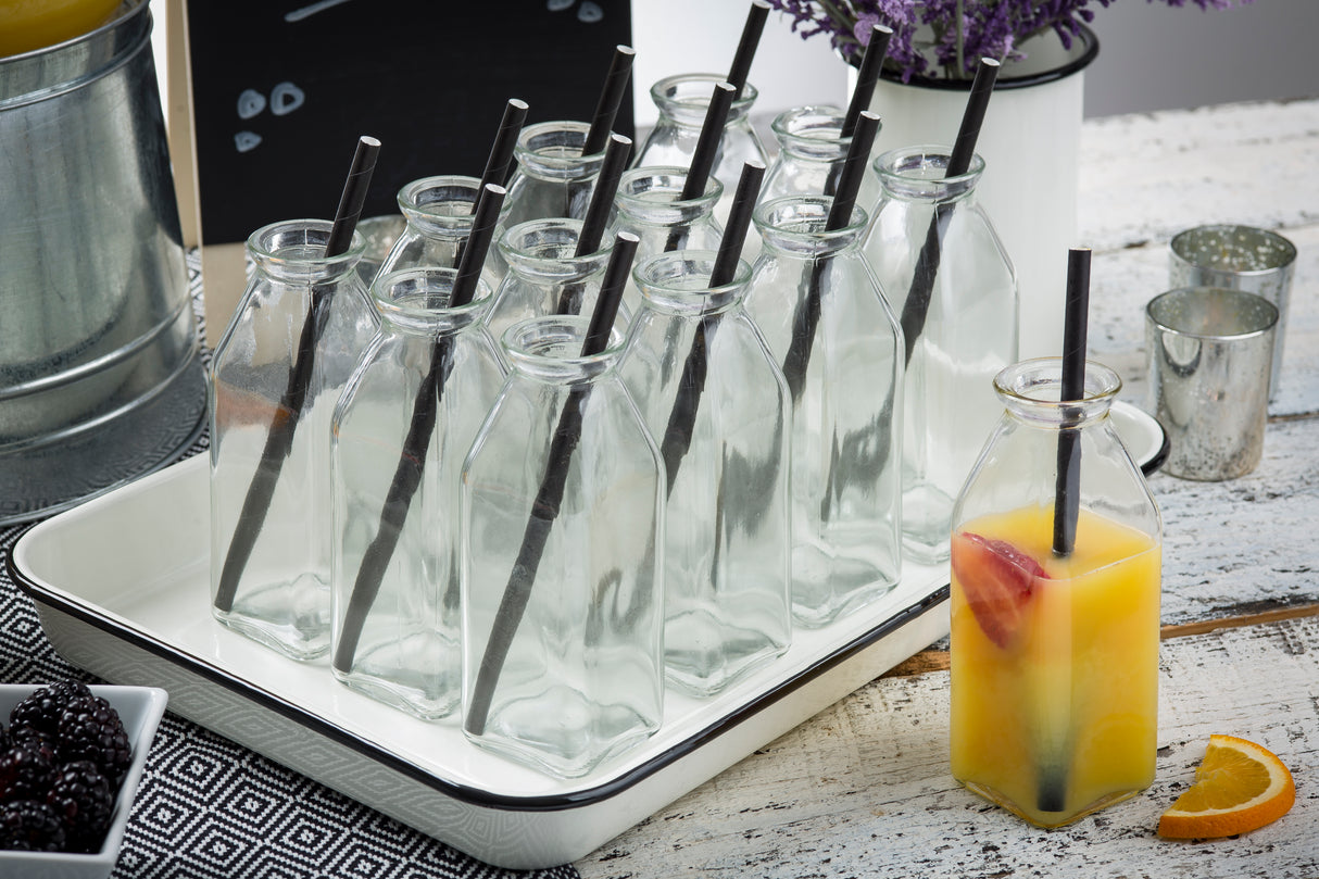 A tray of empty glass milk bottles with black straws sits on a rustic white table. Nearby, a bottle filled with orange juice, fruit slices, and a slice of orange rest on an Enamelware Serving Tray, with blackberries and lavender flowers in the background.