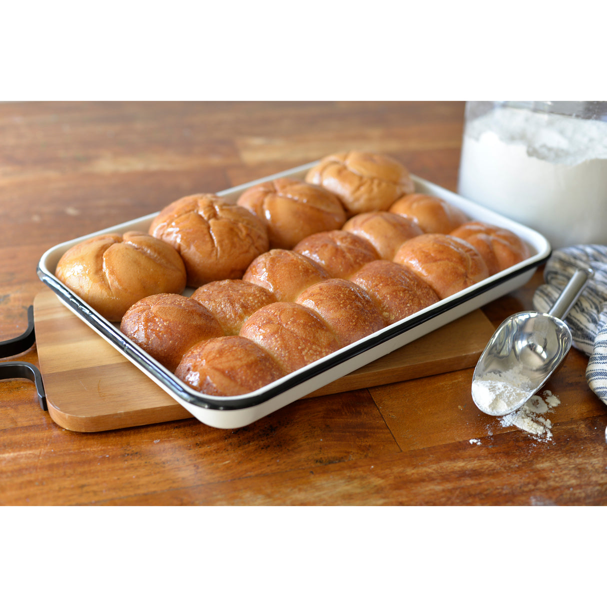 A baking tray filled with shiny, golden-brown dinner rolls sits on a wooden surface. The Porcelain Enamel Tray enhances their appeal, while a metal scoop and jar of flour sit nearby. The closely packed rolls glisten, fresh from the oven.