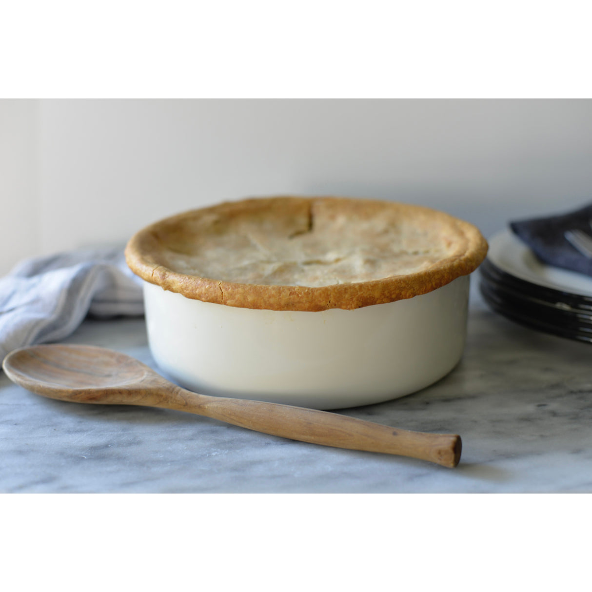 A white porcelain enamel tray with a golden-brown pie crust sits on a marble surface. In front of it is a wooden spoon. A folded dish towel and stacked dark plates with silverware are in the background, against a plain white wall.