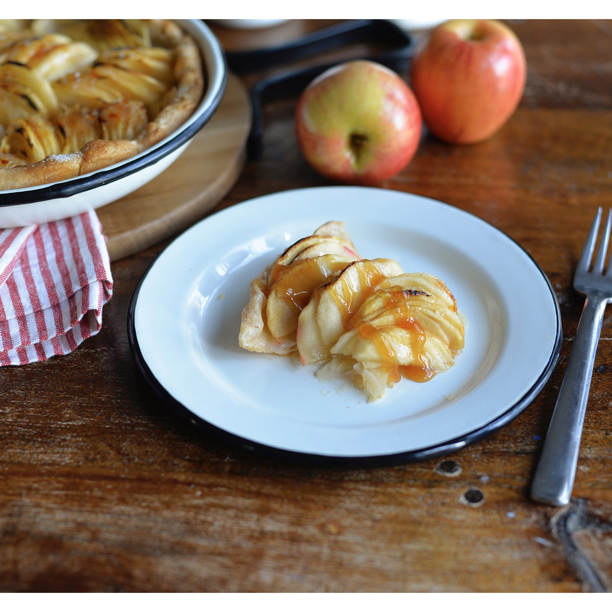 A white enamelware plate holds two slices of glazed apple tart, with golden baked apples on flaky crust. A fork sits to the right. In the background are red apples, a whole tart in a pie dish, and a red-striped napkin on a rustic wooden table.