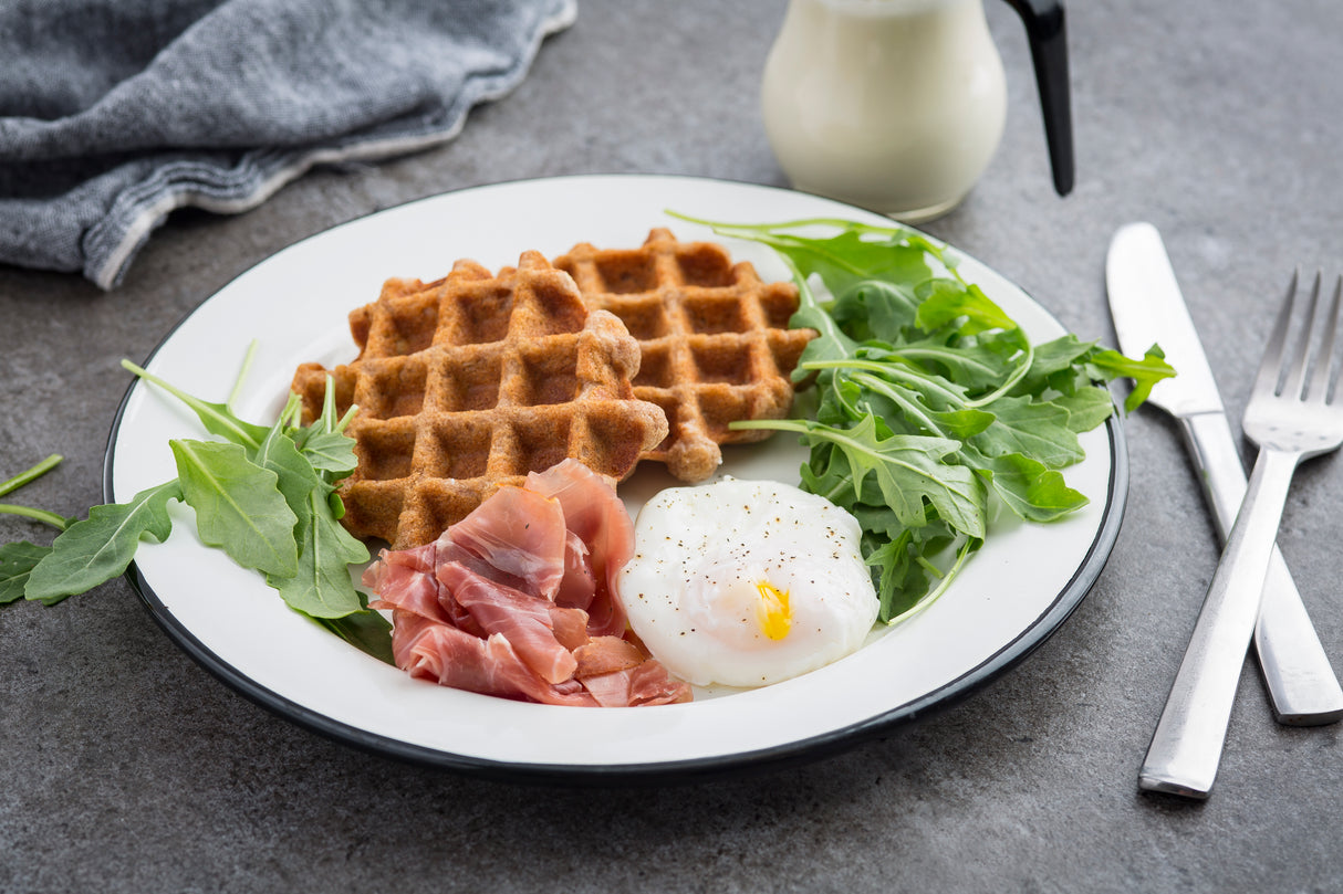 A porcelain enamel plate holds two brown waffles, a poached egg topped with black pepper, prosciutto slices, and fresh arugula. A fork, knife, napkin, and small pitcher of cream rest nearby on the gray tabletop.