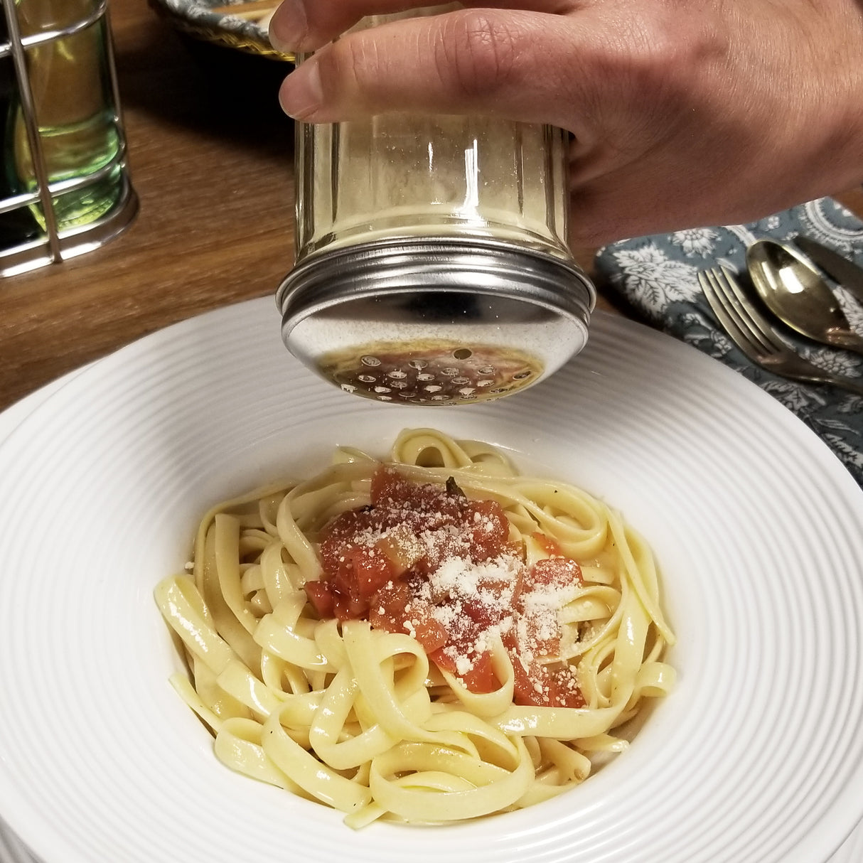 A hand sprinkles grated Parmesan cheese from a stainless steel shaker with a perforated top onto fettuccine pasta topped with tomato sauce. A fork and spoon rest on a patterned napkin, with a glass of water in the background.