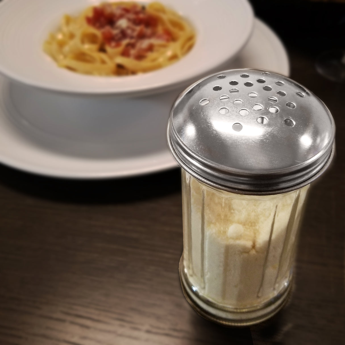 A fluted glass shaker of grated Parmesan cheese with a stainless steel lid sits on a dark wooden table. In the blurred background, a white plate holds pasta with red sauce, possibly carbonara or bolognese, garnished with grated cheese.