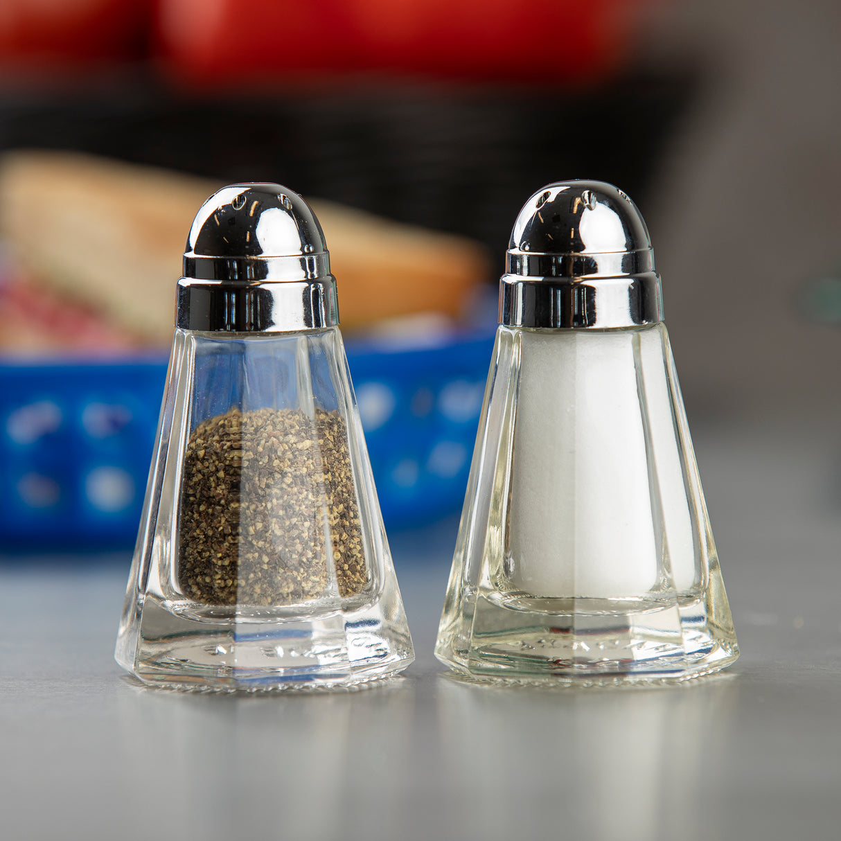 Two glass Salt & Pepper Shakers with stainless steel tops sit side by side; one holds ground black pepper, the other white salt. They rest on a gray surface with a blurred blue basket and red items in the background.