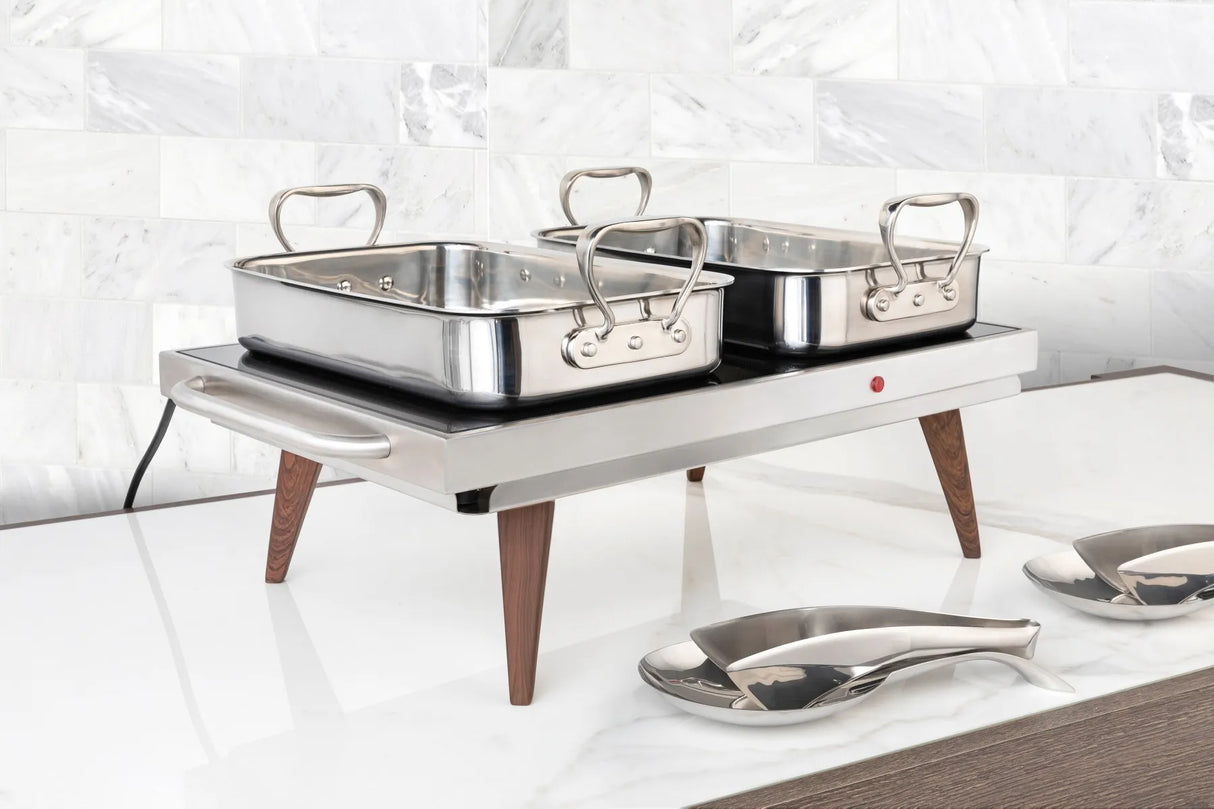 A stainless steel buffet roaster with wooden legs holds two 6 qt chafing dishes. The rectangular warming tray sits on a white marble countertop before a light gray tiled wall, with serving spoons resting on a nearby oval tray.